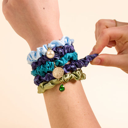 A hand wearing The Darling Effect’s Endless Daydream Navy Scrunchie Charm Set showcases playful navy, purple with a pearl charm, teal with a gold charm, and olive scrunchies as another hand adjusts one accessory.
