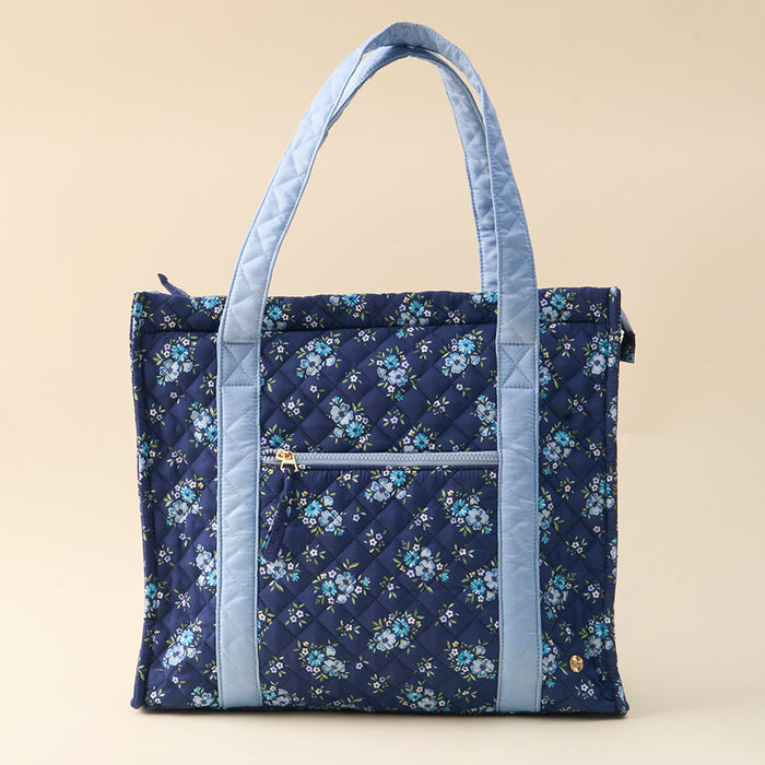 The-darling-effect’s Endless Daydream Navy The Everyday Quilted Tote Bag features a blue floral pattern, light blue handles, and a front zippered pocket—perfect for daily style and convenience, shown against a plain beige background.