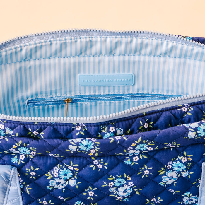 A close-up of the Endless Daydream Navy The Everyday Quilted Tote Bag by the-darling-effect, with blue floral print, light blue and white striped lining, a small zip pocket, and THE DARLING EFFECT label—ideal for daily use.