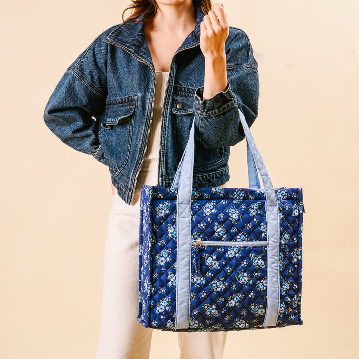 A woman in a denim jacket holds the-darling-effect’s Endless Daydream Navy The Everyday Quilted Tote Bag—a chic blue floral tote ideal for daily use or stylish travel.