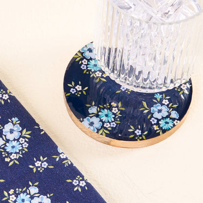 A glass rests on the-darling-effect’s Endless Daydream Navy Wood Enamel Coaster, featuring blue and white florals. To the left, a navy fabric with matching patterns complements this stylish and durable drink coaster.