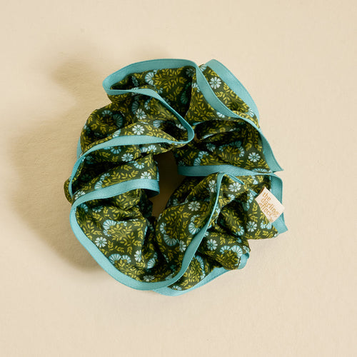 The Darling Effect's Everbloom Forest Green Ruffle Up Scrunchie, featuring green and blue florals with light blue trim and a small fabric tag, is shown on a beige background.