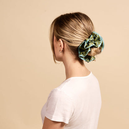 A woman with blonde hair in a low bun, secured with The Darling Effect's Everbloom Forest Green Ruffle Up Scrunchie, wears a white t-shirt and small hoop earrings as she faces away from the camera against a beige background.