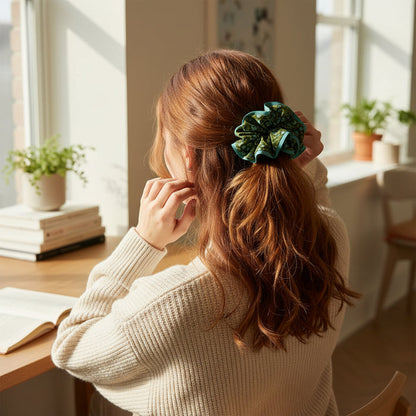 A woman with long, wavy red hair, wearing a cream sweater and The Darling Effect's Everbloom Forest Green Ruffle Up Scrunchie, sits at a wooden desk by a window with potted plants and an open book in a sunlit room.