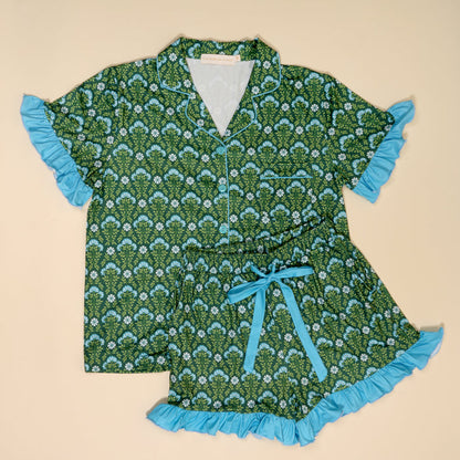 The Darling Effect’s Everbloom Forest Green Slumber Party Pajama Shorts Set features ultra-soft fabric, a light blue floral pattern, blue trim and ruffles, a button-up top with pocket, and shorts with a blue ribbon waist tie.