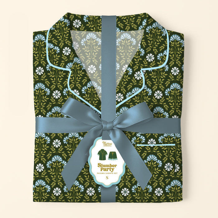 A folded Everbloom Forest Green Slumber Party Pajama Shorts Set by The Darling Effect, featuring a blue and white floral print, is tied with a ribbon. A tag displays an illustration and the set’s name.
