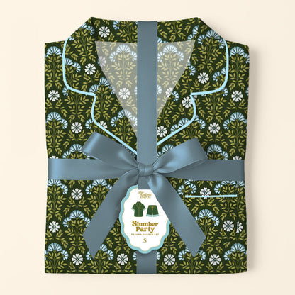 A folded Everbloom Forest Green Slumber Party Pajama Shorts Set by The Darling Effect, featuring a blue and white floral print, is tied with a ribbon. A tag displays an illustration and the set’s name.