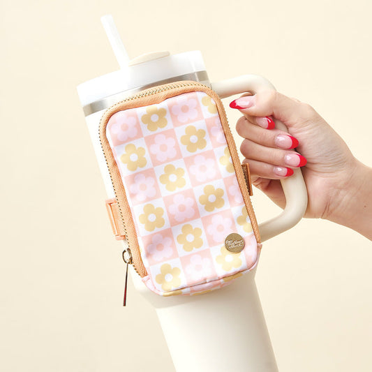 A hand with pink and white manicured nails holds a large white tumbler with a beige straw, featuring The Darling Effect’s Flower Check Peach Tumbler Fanny Pack—a water-resistant accessory with a pink, yellow, and white checkered flower pattern.