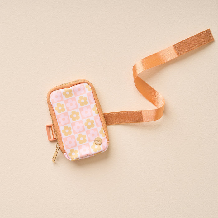 The Darling Effect's Flower Check Peach Tumbler Fanny Pack—a small, rectangular pouch with a pink and yellow retro floral pattern, gold zipper, tan trim, and adjustable peach strap—lies flat on a light beige surface.