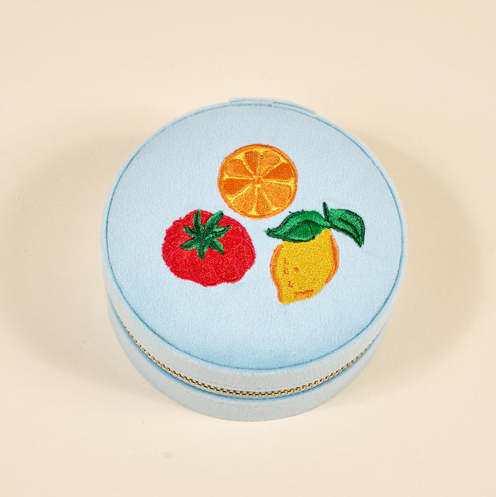 The Darling Effect Fruit Market Round Velvet Jewelry Case is a light blue, gold-zippered box with embroidered orange, lemon, and tomato designs—an ideal travel organizer for your favorite accessories.