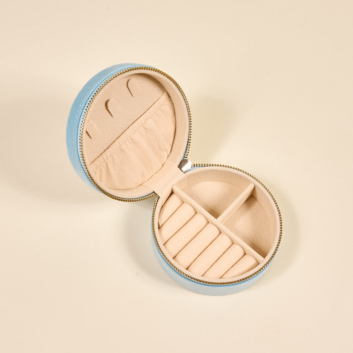 The Darling Effect’s Fruit Market Round Velvet Jewelry Case in light blue is open on a plain surface, showing a beige interior with ring slots, small compartments, and a pocket to organize jewelry.