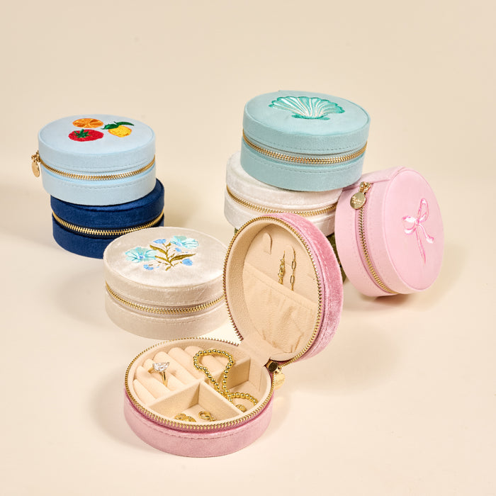 Six Fruit Market Round Velvet Jewelry Cases by The Darling Effect in pastel hues feature unique embroidered designs and gold zippers. The open pink organizer displays jewelry, while the other cases are closed and stacked behind.