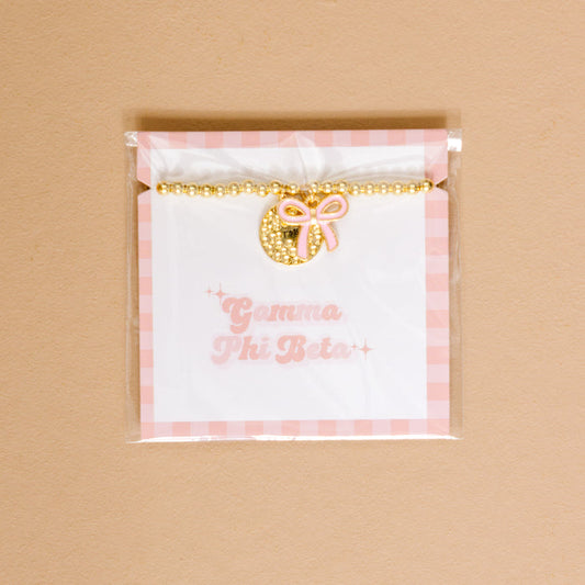 The Darling Effect’s Gamma Phi Beta Bow Gold Charm Bracelet, featuring gold beads, a pink bow, and a circular charm, is displayed on a white and pink gingham card—perfect for showing sorority pride against a chic beige background.