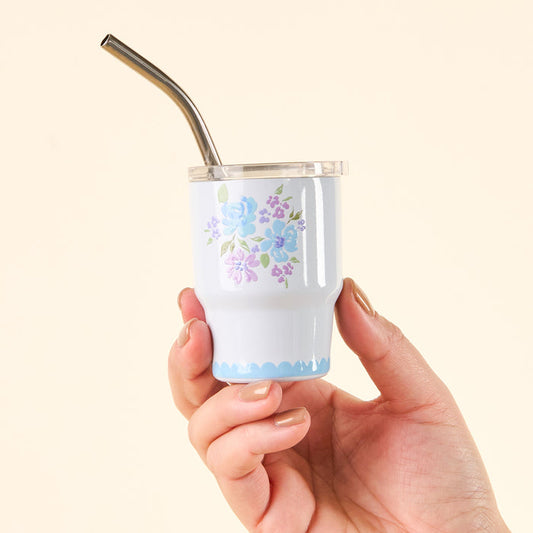 A hand holds the-darling-effect’s Garden Blooms Blue 2 oz Tiny Tumbler featuring a floral design, clear lid, and metal straw against a light beige background.