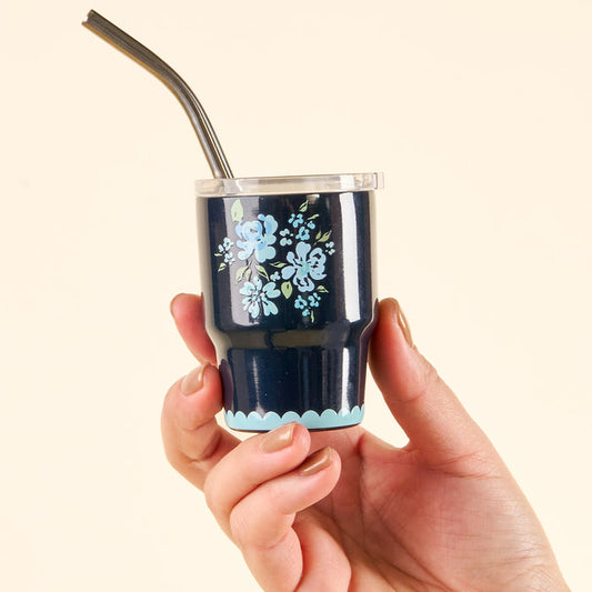 A hand holds the-darling-effect’s Garden Blooms Navy 2 oz Tiny Tumbler—a small navy cup featuring light blue floral designs, a clear lid, and a metal straw—set against a plain light background.