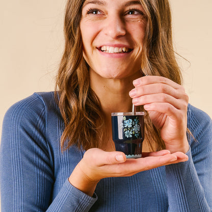 A smiling woman in a blue top cheerfully holds and stirs the-darling-effect's Garden Blooms Navy 2 oz Tiny Tumbler filled with a dark drink.