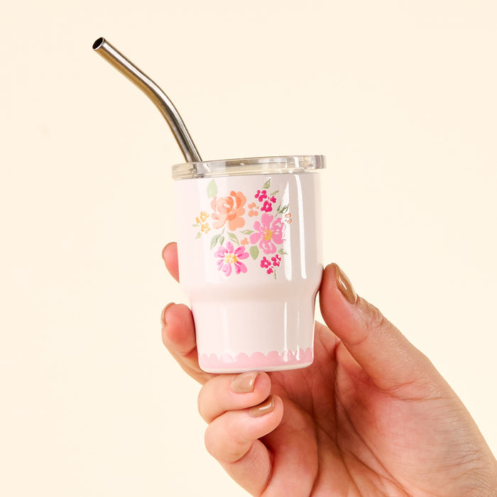 A hand holds the-darling-effect’s Garden Blooms Pink 2 oz Tiny Tumbler, featuring cute prints, a clear lid, and a metal straw against a light beige background.