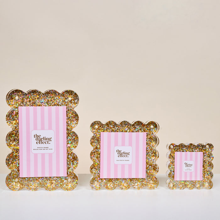 Three Gold Glitter Rectangle Scalloped Acrylic Picture Frames by The Darling Effect in graduated sizes are displayed with pink striped insert cards on a white surface against a neutral background.
