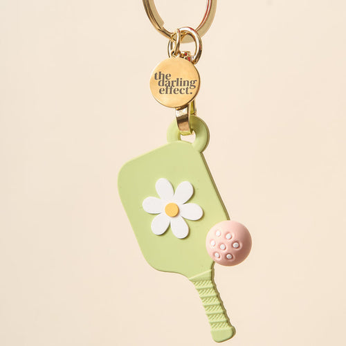 The Darling Effect’s Green Pickleball Paddle Keychain features a light green paddle with a white daisy, a pink ball, and a gold keyring with an engraved charm—making it the perfect pickleball gift.