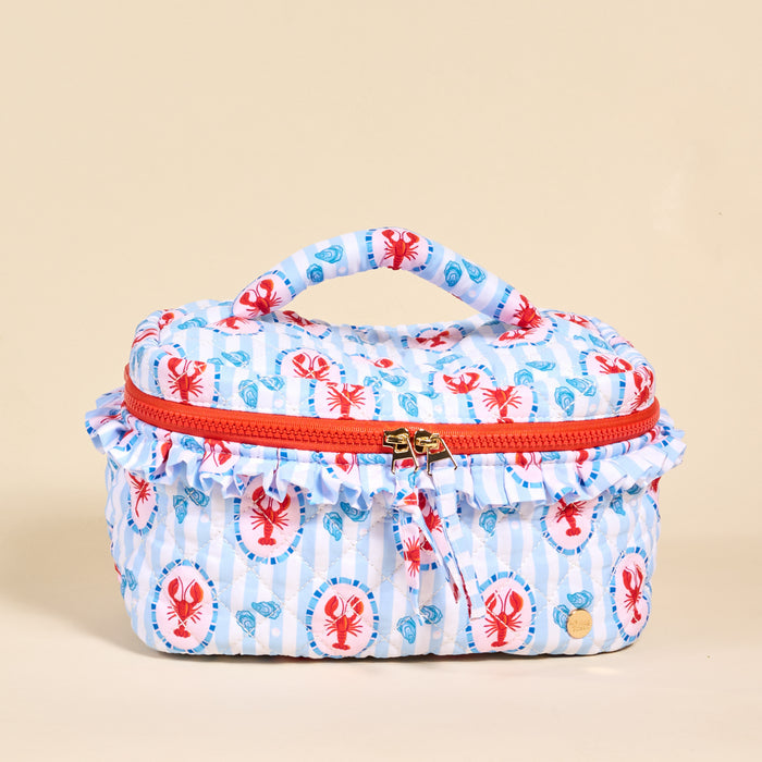 The Darling Effect’s Harbor House Glam & Go Quilted Cosmetic Bag is a rectangular bag with a top handle, red zipper, and ruffled trim, featuring red lobsters and blue seashells on white and blue stripes.