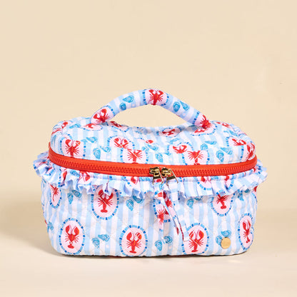 The Darling Effect’s Harbor House Glam & Go Quilted Cosmetic Bag is a rectangular bag with a top handle, red zipper, and ruffled trim, featuring red lobsters and blue seashells on white and blue stripes.