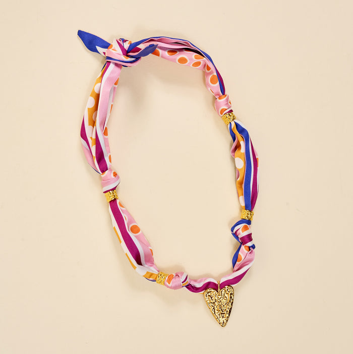 The Darling Effect’s Heart of Gold Make a Statement Scarf Necklace features pink, purple, and orange silk stripes and dots, gold beads, and a textured gold heart pendant for an artful look on beige.