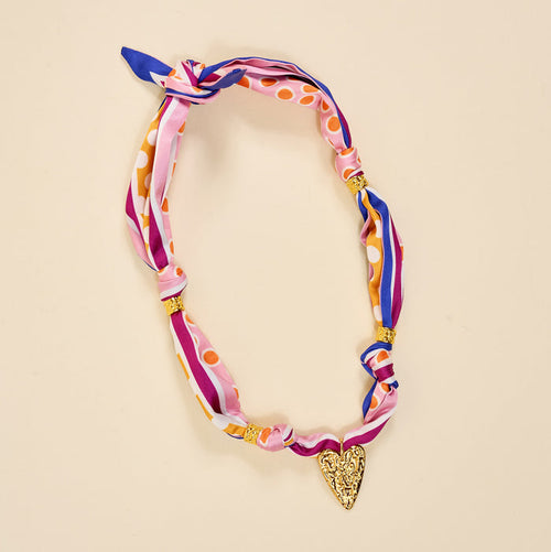 The Darling Effect’s Heart of Gold Make a Statement Scarf Necklace features pink, purple, and orange silk stripes and dots, gold beads, and a textured gold heart pendant for an artful look on beige.