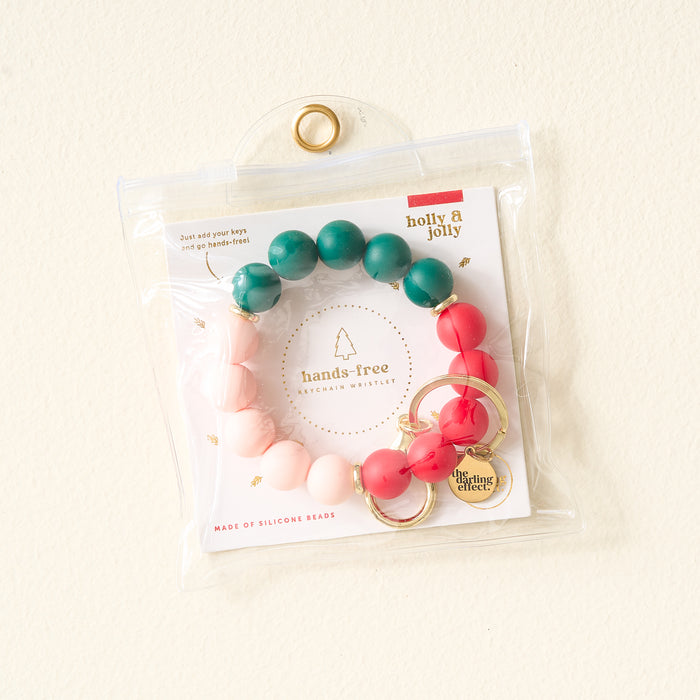 The Darling Effect’s Holly Jolly Hands-Free Keychain Wristlet features green, red, and pink silicone beads with gold hardware and a charm, hanging on a white wall.