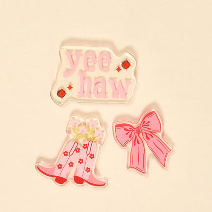 The Darling Effect's Howdy Honey Little Magnet Set features three colorful accents: a "yee haw" design in pink with red diamonds, a pair of pink cowboy boots with flowers, and a large pink bow, all on a beige background.