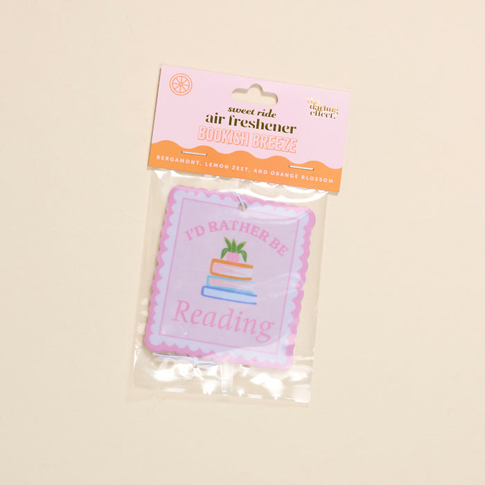 The Darling Effect’s “I’d Rather Be Reading Sweet Ride” car air freshener features bergamot, lemon zest, and orange blossom scents, plus a book stack illustration and the phrase “I’d Rather Be Reading.”.
