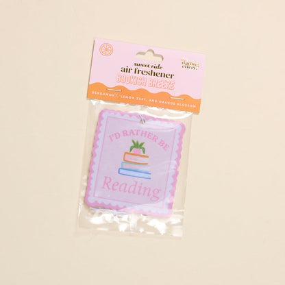 The Darling Effect’s “I’d Rather Be Reading Sweet Ride” car air freshener features bergamot, lemon zest, and orange blossom scents, plus a book stack illustration and the phrase “I’d Rather Be Reading.”.