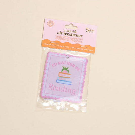 The Darling Effect’s “I’d Rather Be Reading Sweet Ride” car air freshener features bergamot, lemon zest, and orange blossom scents, plus a book stack illustration and the phrase “I’d Rather Be Reading.”.