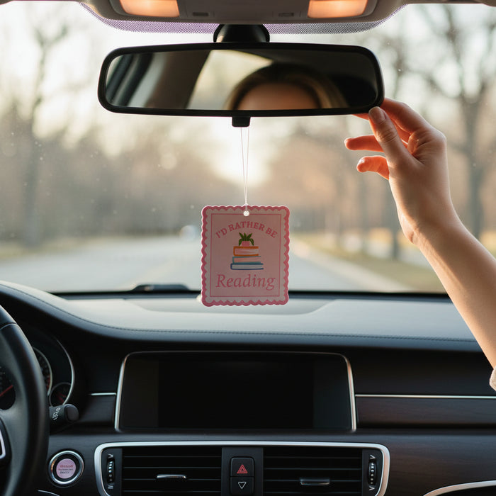 A person in a car adjusts The Darling Effect's I'd Rather Be Reading Sweet Ride Car Air Freshener, featuring stacked books and hanging from the rearview mirror. The scent fills the air as the dashboard and steering wheel are visible.