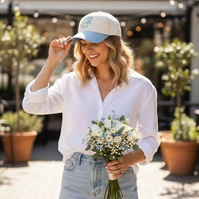 A bride in a white linen shirt and denim shorts adjusts her In My Bride Era Classic Canvas Hat by The Darling Effect, holding a white and blue bouquet—a laid-back look for a casual celebration.