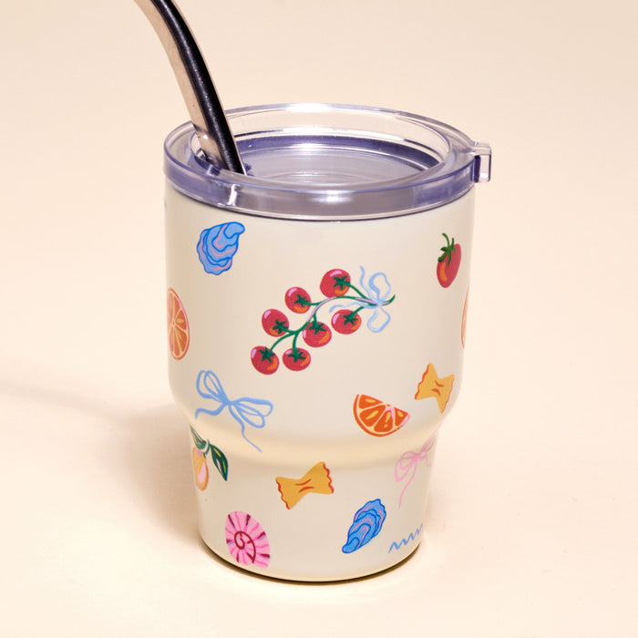 The Darling Effect's Italian Summer 2 oz Tiny Tumbler features a cream-colored design with a clear lid, metal straw, and vibrant illustrations of tomatoes, oranges, candy, bows, strawberries, and seashells on a light beige background.