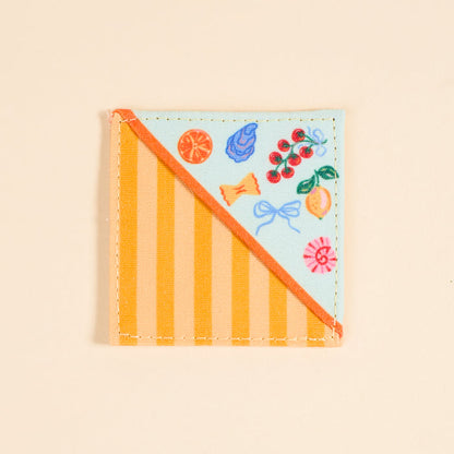 The Darling Effect’s Italian Summer Corner Keeper Fabric Bookmark features diagonal orange stripes on one side and vibrant fruit, candy, and ribbon illustrations on the other—a perfect gift for book lovers. Set against a light beige background.
