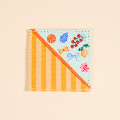 The Darling Effect’s Italian Summer Corner Keeper Fabric Bookmark features diagonal orange stripes on one side and vibrant fruit, candy, and ribbon illustrations on the other—a perfect gift for book lovers. Set against a light beige background.