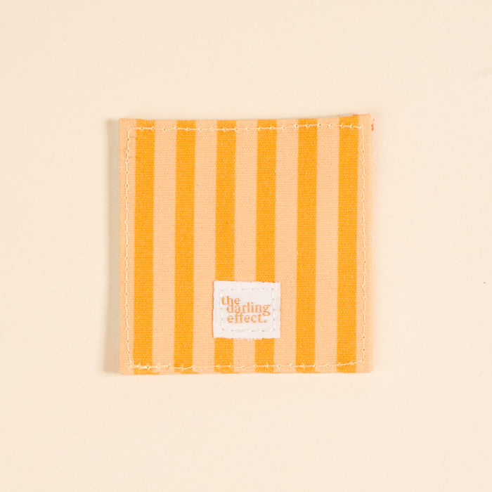 A square fabric coaster with vertical yellow and light beige stripes, white stitching, and a small "The Darling Effect" label. Perfect paired with the Italian Summer Corner Keeper Fabric Bookmark or as a thoughtful gift for book lovers.