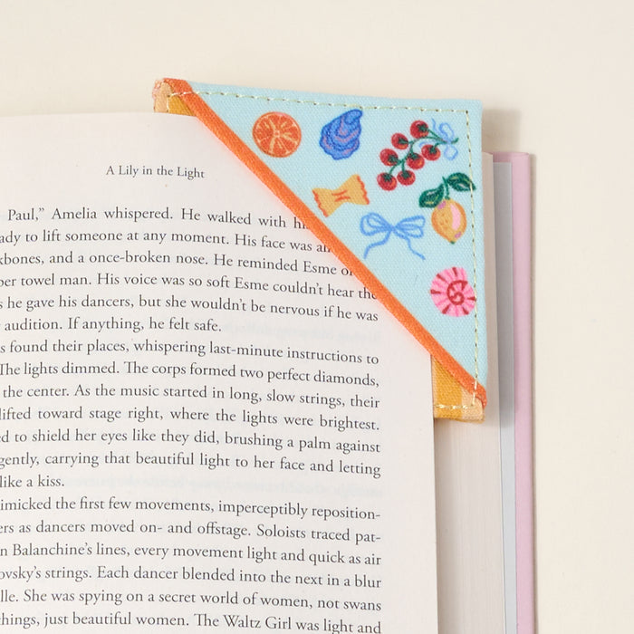 The Darling Effect's Italian Summer Corner Keeper Fabric Bookmark set includes two bookmarks—one orange with fruit and dessert designs, one light purple—making a unique book lover gift. Shown resting on an open book against a neutral background.