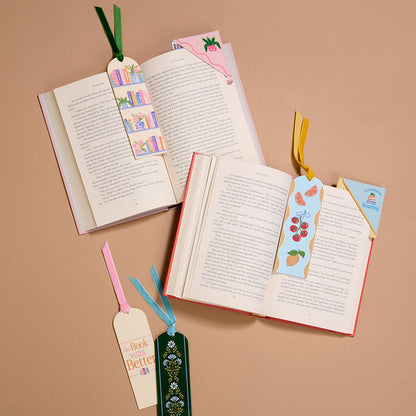Three open books rest on a beige surface, each marked with an Italian Summer Corner Keeper Fabric Bookmark by The Darling Effect. Two more uniquely designed bookmarks with ribbon ties, perfect for book lovers, are displayed beside the books.