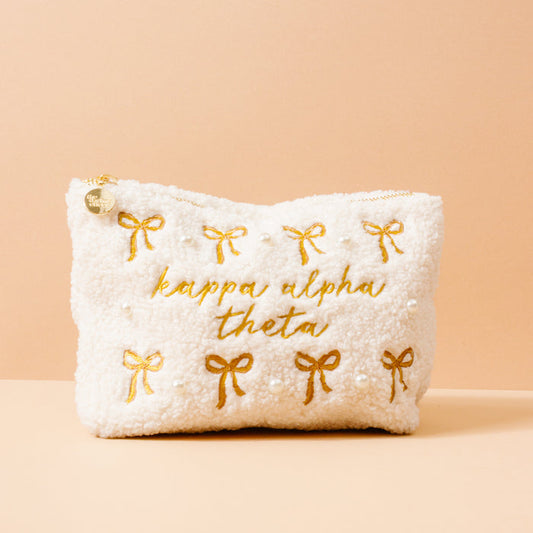 The Darling Effect’s Kappa Alpha Theta Bows & Pearls Teddy Pouch features cream sherpa, a gold zipper, embroidered bows, pearl accents, and “kappa alpha theta” in gold—an ideal sorority gift.