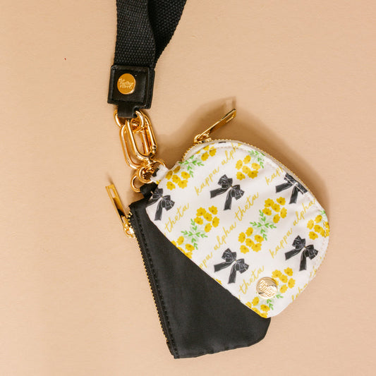 The Darling Effect Kappa Alpha Theta Dynamic Duo Pouch Wristlet features floral patterns and “happy alpha theta,” attached to a black lanyard with gold hardware and zippered pouches, all hands-free on a beige surface.