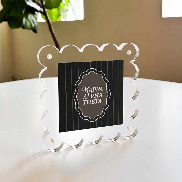 The Darling Effect's Kappa Alpha Theta Square Scalloped Acrylic Frame features white text on a black center. The clear frame stands on a white table by a window with natural light and a leafy plant in the background.
