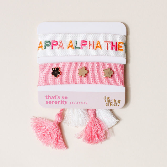 The Darling Effect’s Kappa Alpha Theta Woven Bracelet Set includes a white bracelet with “KAPPA ALPHA THETA” in rainbow letters and a pink band with three gold flower charms, finished with pink and white tassels.