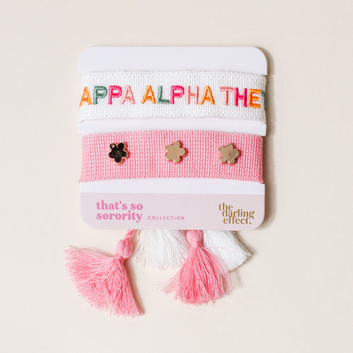 The Darling Effect’s Kappa Alpha Theta Woven Bracelet Set includes a white bracelet with “KAPPA ALPHA THETA” in rainbow letters and a pink band with three gold flower charms, finished with pink and white tassels.