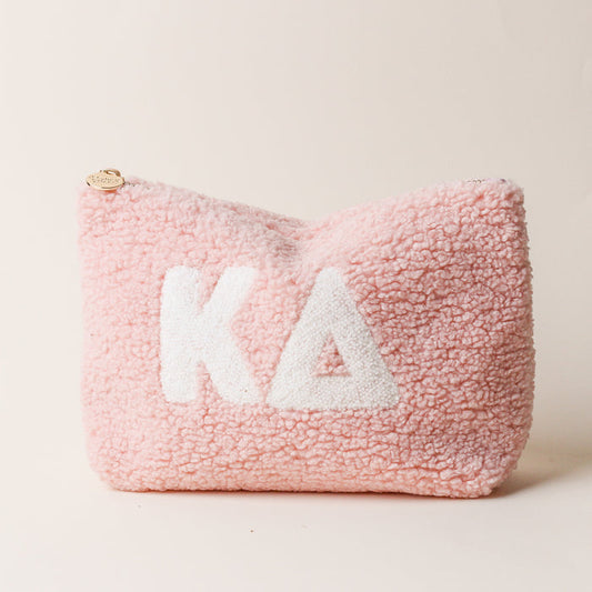 The Darling Effect’s Kappa Delta Blush Teddy Letter Pouch is a soft, pink sherpa pouch with white KΔ Greek letters and a small round gold zipper pull—perfect as a carry-on accessory, shown on a plain light background.