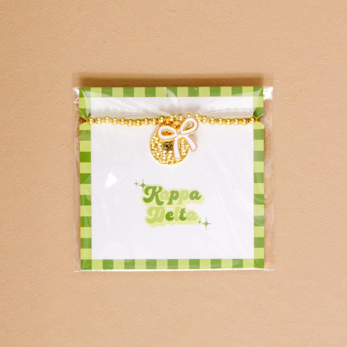 The Darling Effect's Kappa Delta Bow Gold Charm Bracelet features a lemon and bow charm on a white card with green checkered borders and Kappa Delta in green script, all in clear packaging—perfect for showing your chapter pride.