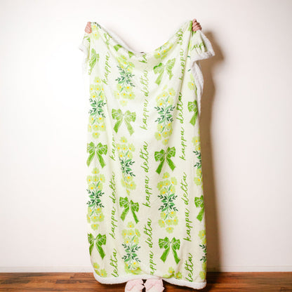 A person holds The Darling Effect’s Kappa Delta Plush Sherpa Blanket, featuring green bows, yellow flowers, and "happy" in green script—an ideal cozy throw for any college dorm.