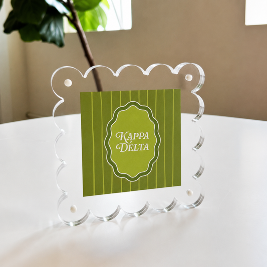 The Darling Effect Kappa Delta Square Scalloped Acrylic Frame displays a green striped card with white "Kappa Delta" text on a round table, set against a background of leafy plant and window.