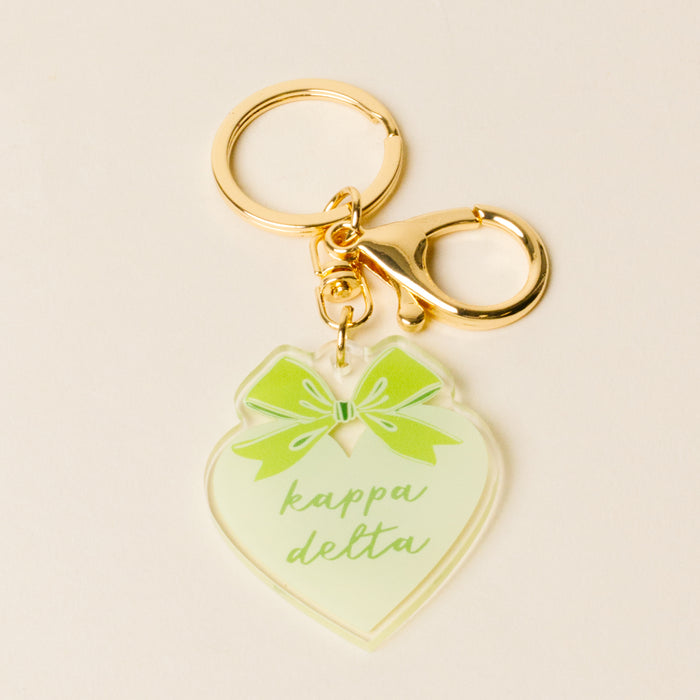 The Darling Effect's Kappa Delta Sweetheart Bow Acrylic Keychain features a gold heart, green bow, and "kappa delta" in light green script—a perfect sisterhood gift for any sorority member.